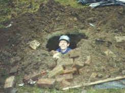 a man in a hard hat peeks out from a deep hole dug into dirt and scattered bricks