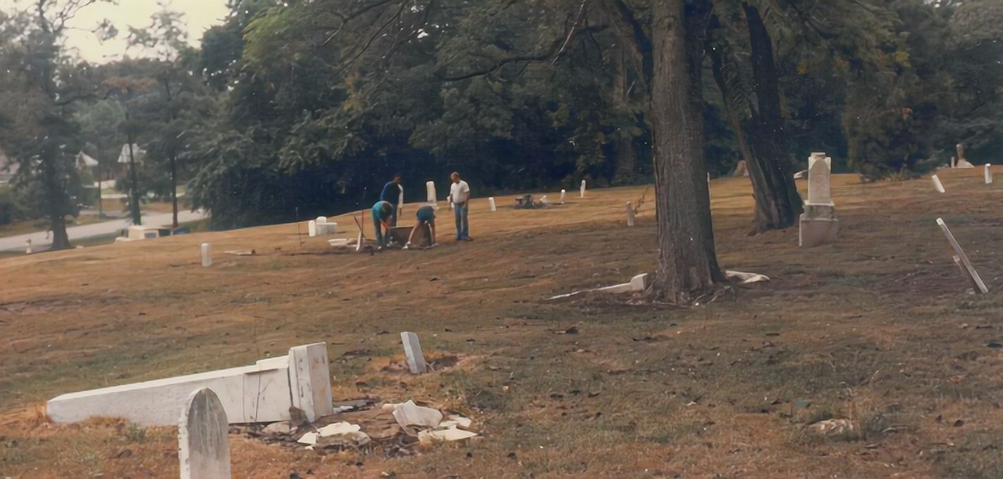 four people lift a stone grave marker off the ground and are surrounded by many more upright and fallen white and grey tombstones and old trees in a sloping grassy cemetery